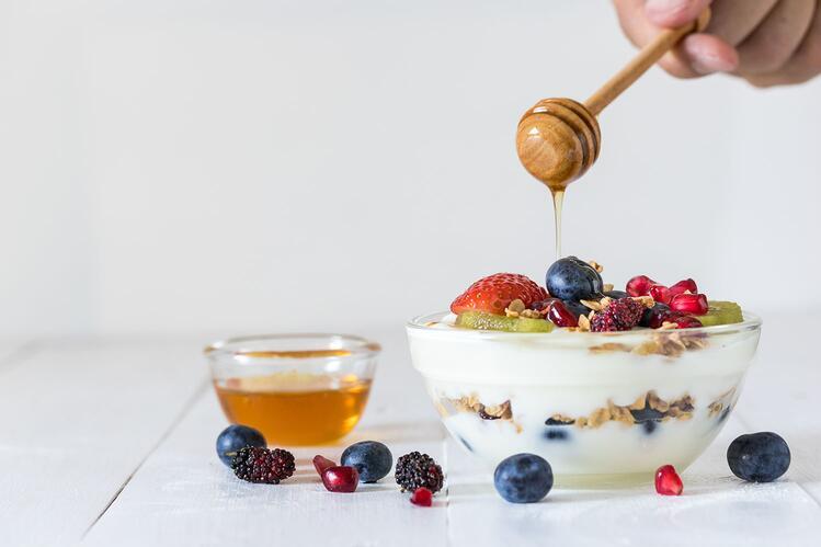 A bowl of yogurt parfait on a table with honey dripping into it, surrounded by a smaller bowl of honey and fresh fruit. 