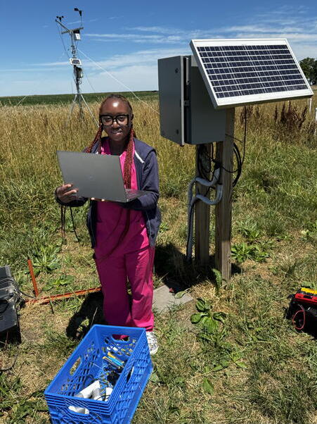 Kyeaira Faustin in the field during her time in the Center for Digital Agriculture’s summer program Kyeaira Faustin stands outdoors with a laptop near a solar panel, electrical box, and weather monitoring equipment, surrounded by tall grass on a sunny day